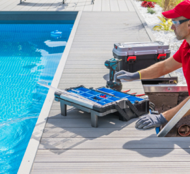 Outdoor Swimming Pool Heating and Filtering System Maintenance Performed by Caucasian Middle Aged Technician. Open Toolbox and Electric Drill Driver on the Floor in Front of Him.